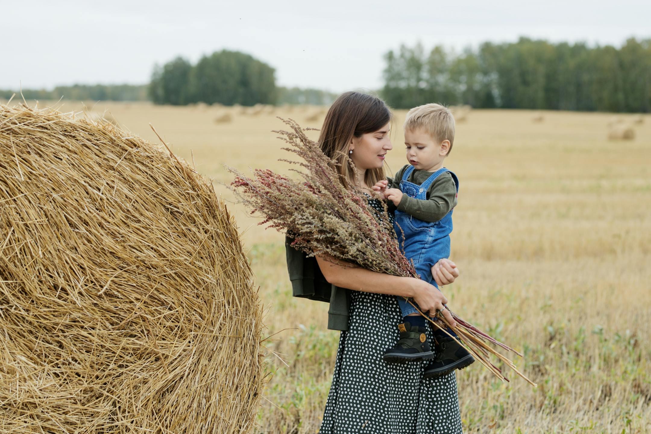 A mother holding her child and bouquet of wildflowers in a hayfield on a bright day.