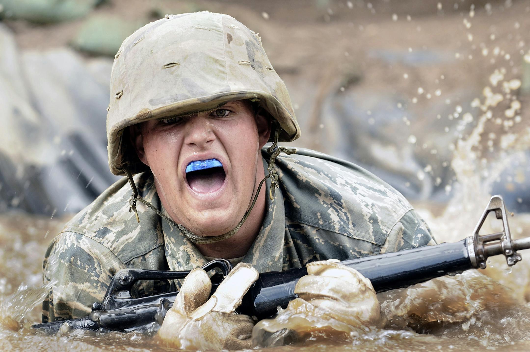 Soldier in military training crawling through muddy water with a rifle and mouthguard.
