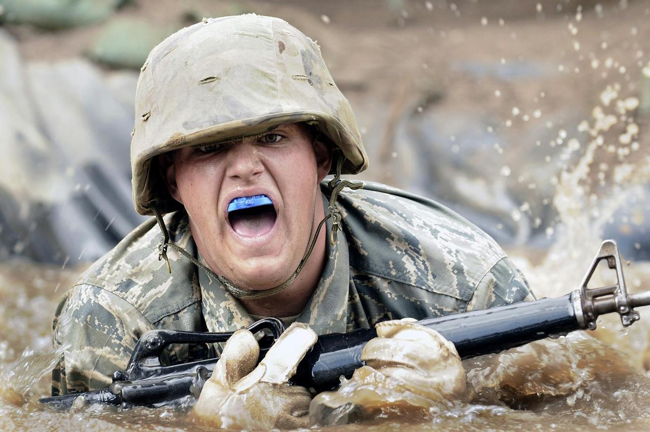 Soldier in military training crawling through muddy water with a rifle and mouthguard.