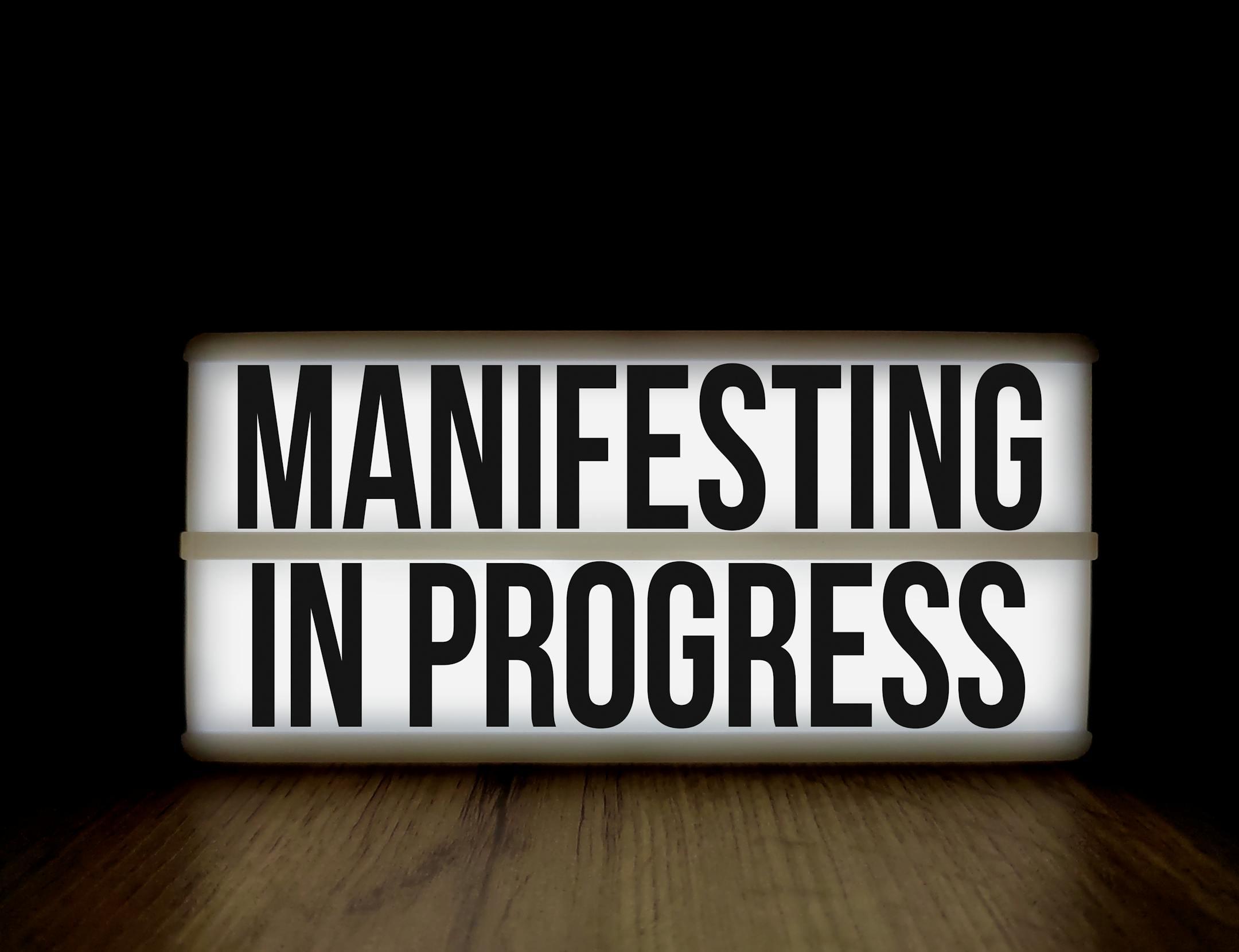 Illuminated lightbox sign with the motivational message 'Manifesting in Progress'.