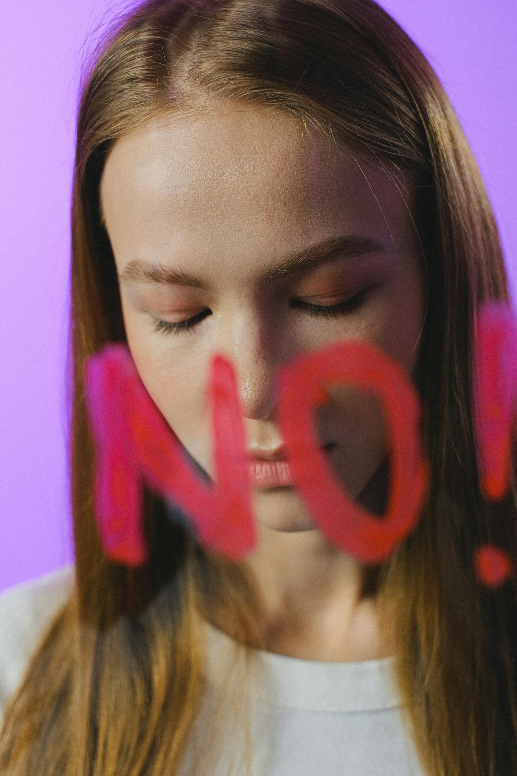 A teenager with brown hair reflecting, with 'No' written on glass, set against a vibrant pink background.