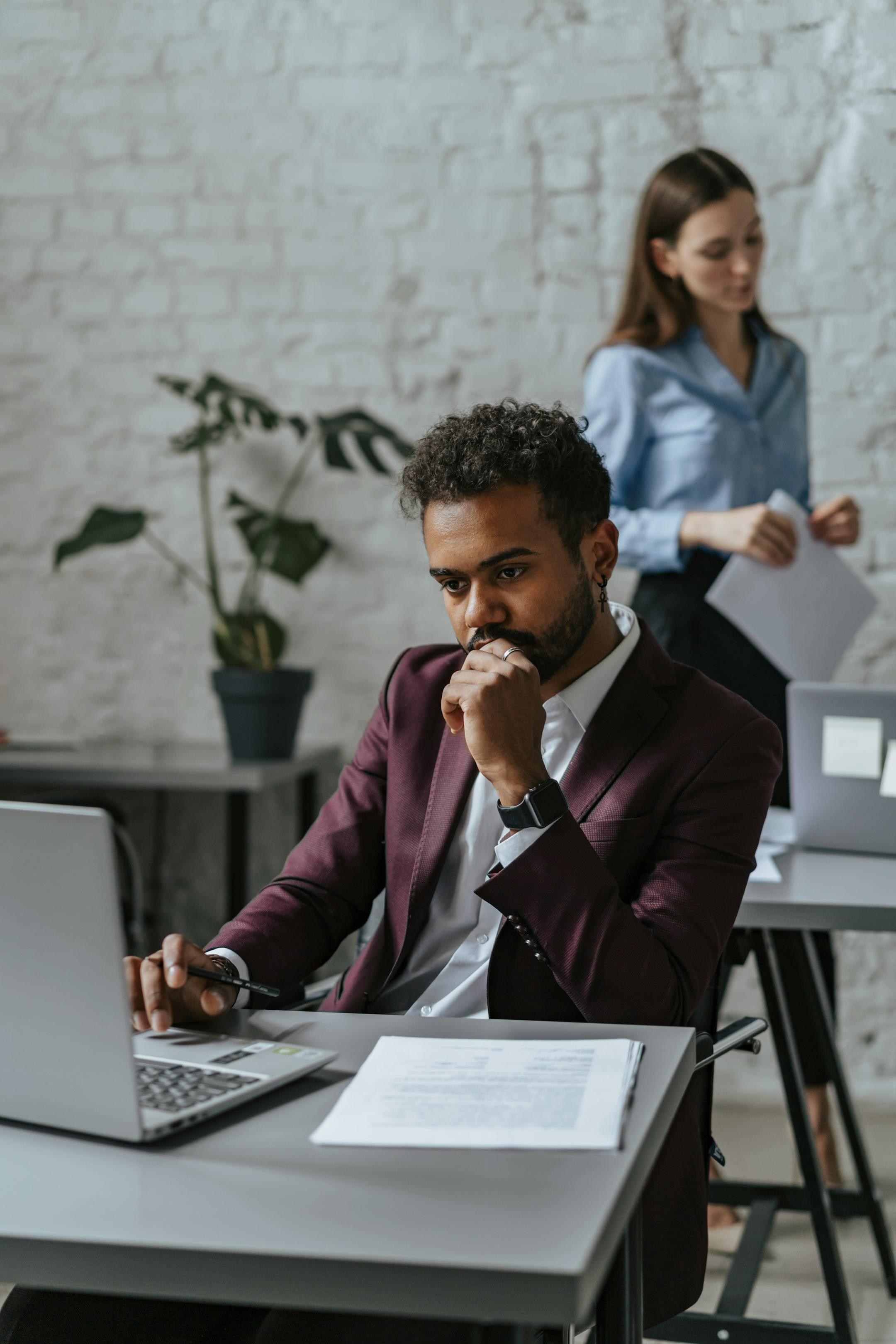 Pensive man using laptop in modern office with female colleague in background.
