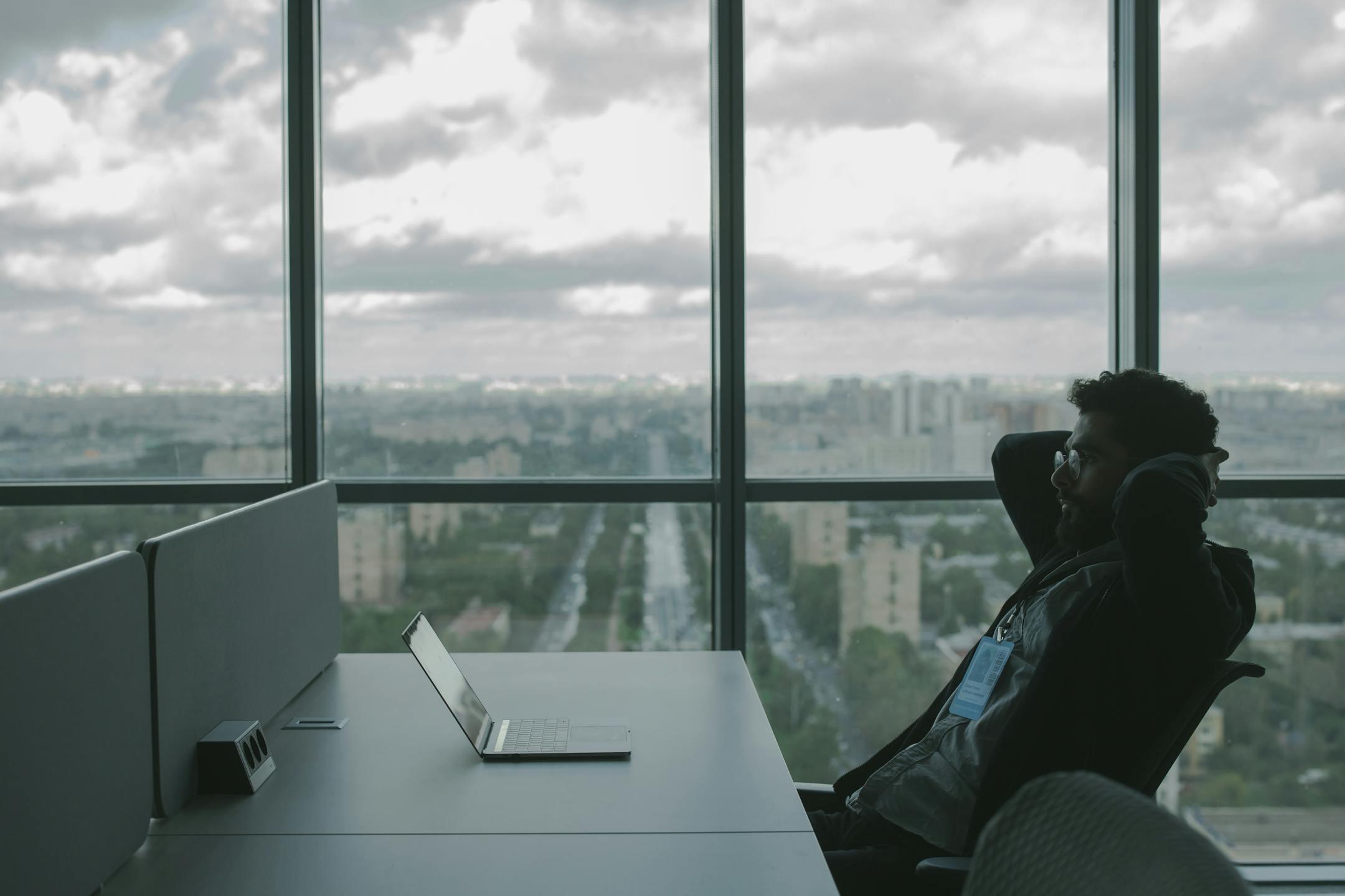 A man sits in an office with hands on head in front of a laptop, overlooking a cityscape.