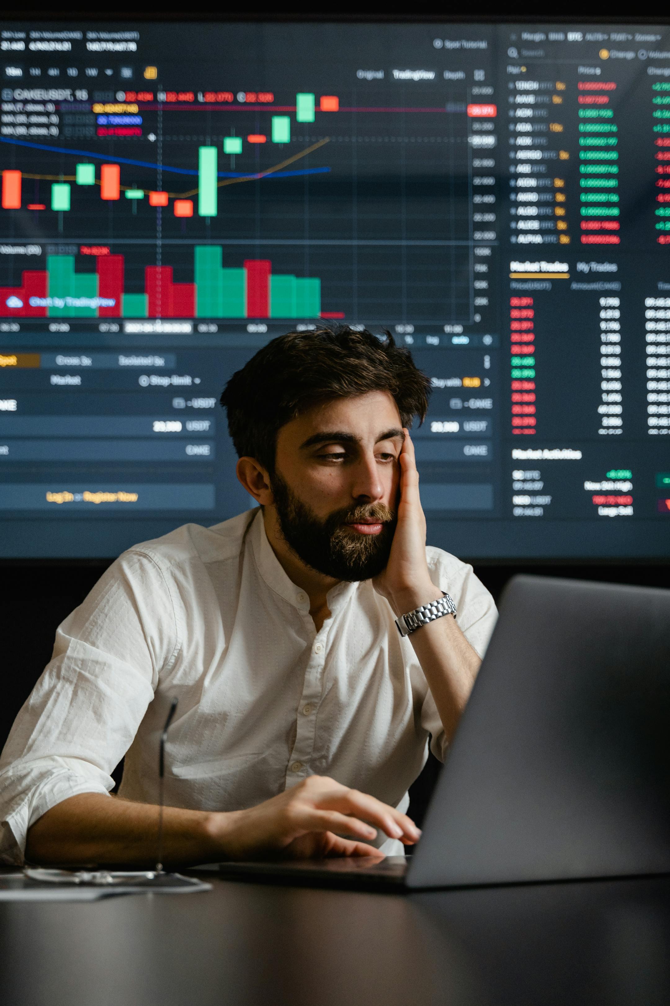 Bearded businessman analyzing financial statistics on a laptop, with stock market graphs in the background.