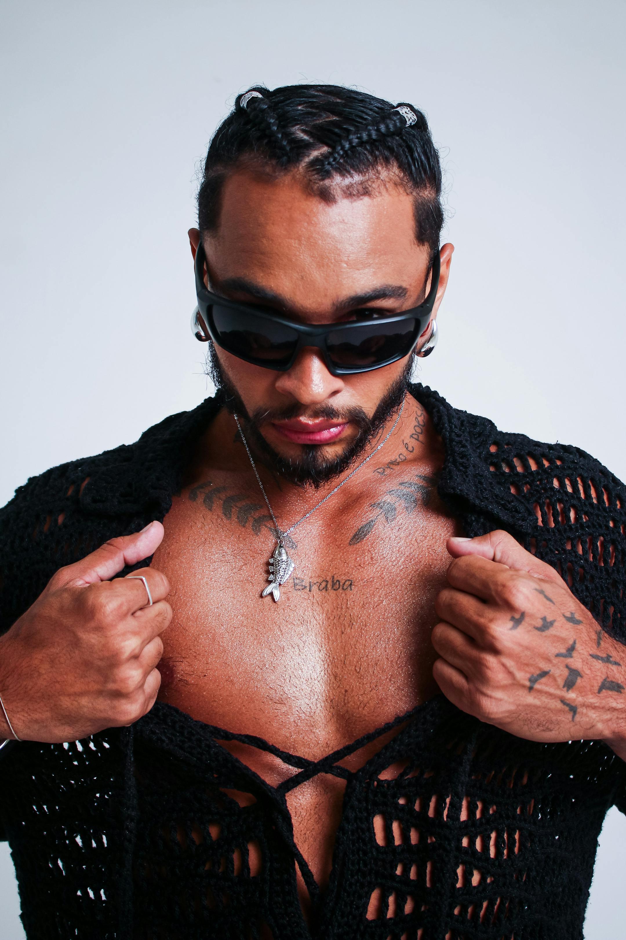 Confident man in sunglasses and tattoos posing with open shirt.