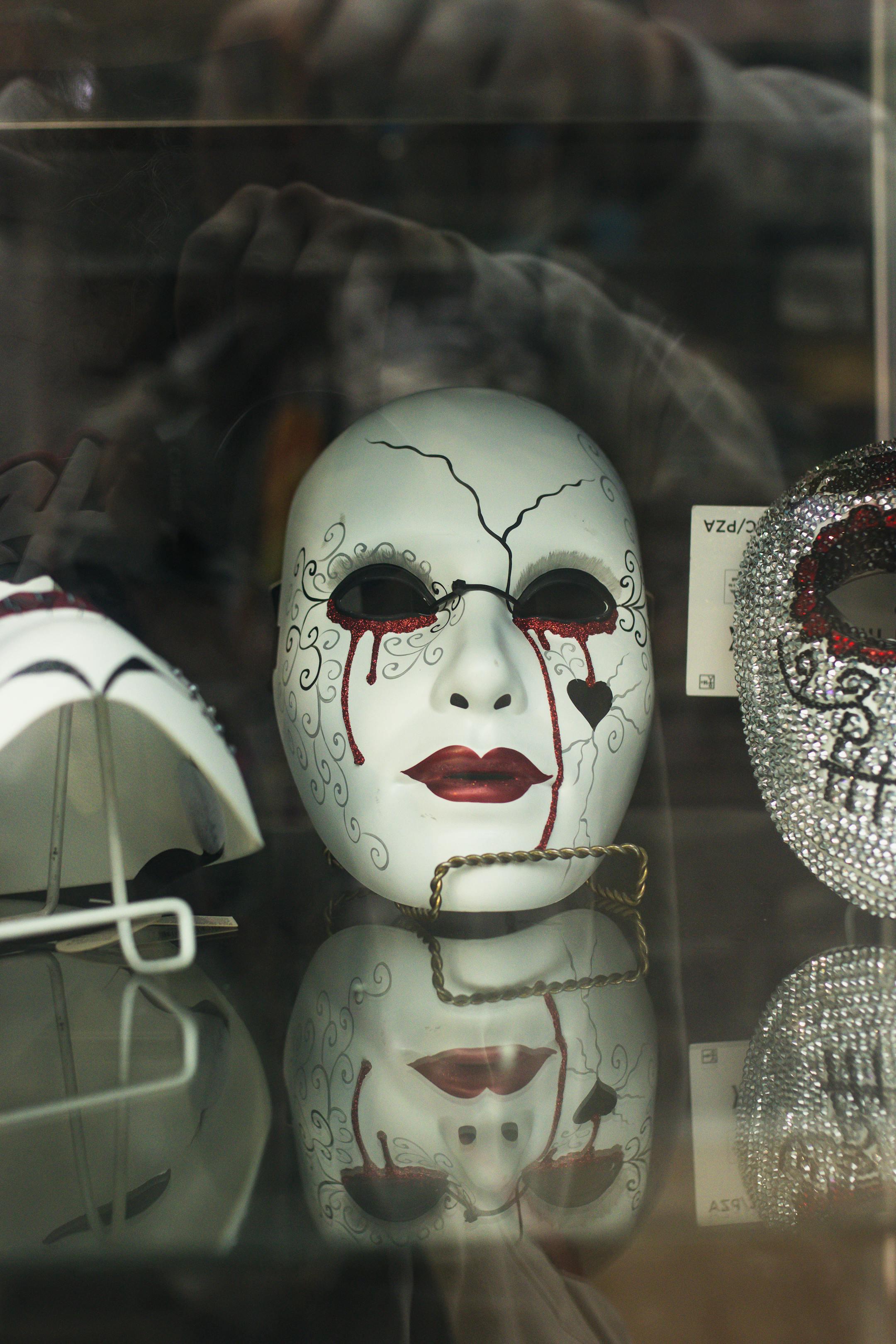 A dramatic artistic mask with red markings displayed in a reflective glass case.