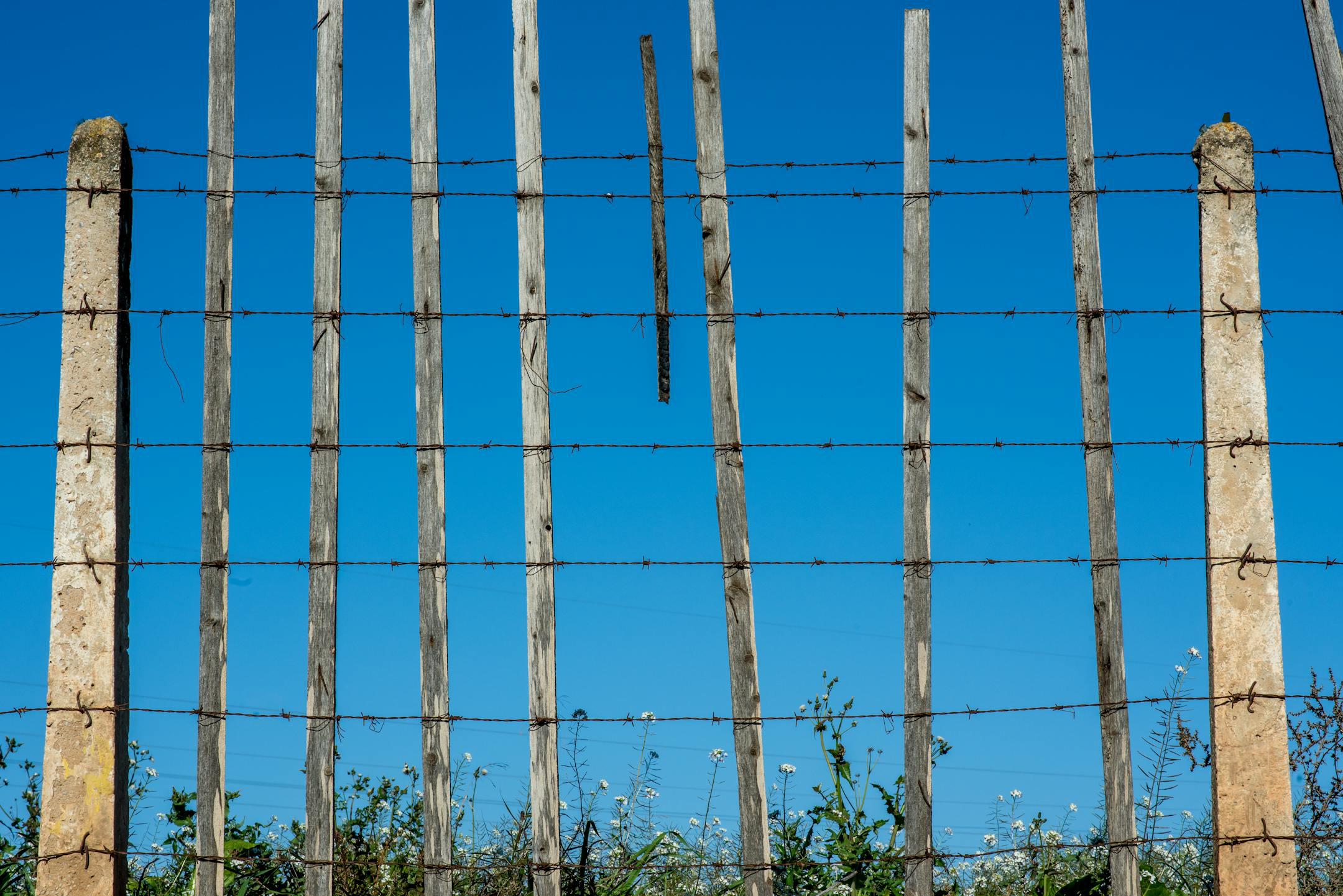 Rustic wooden and concrete fence with barbed wire against a clear blue sky.
