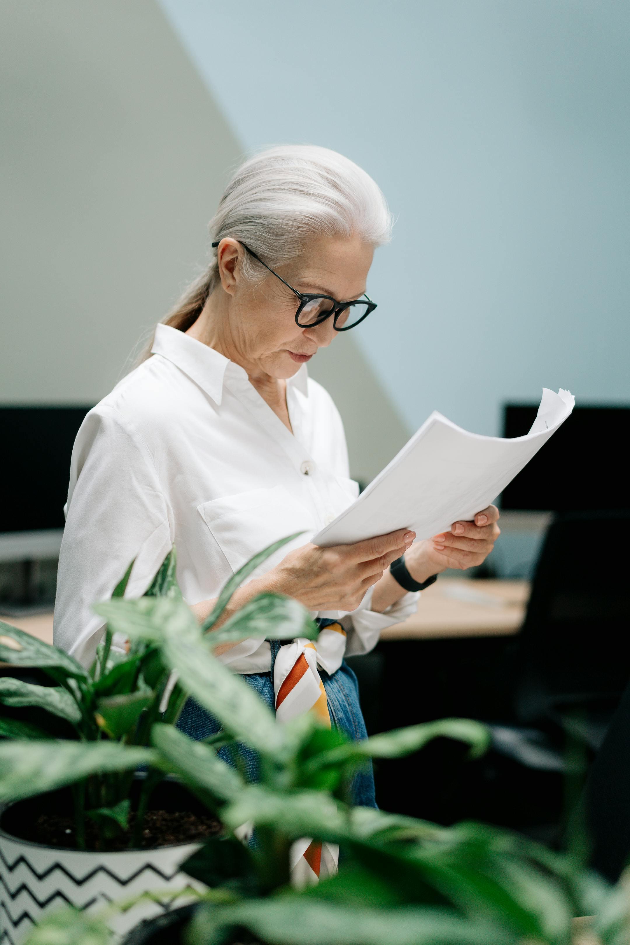 Elderly woman with eyeglasses reviews papers in modern office, surrounded by plants.